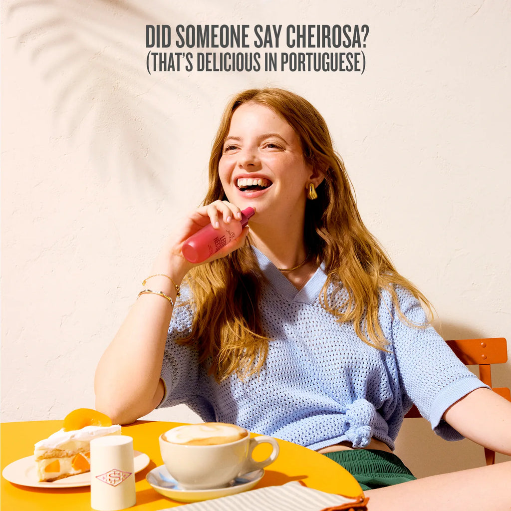 Woman sitting at a table with a cup of coffee and a pastry, laughing and holding a pink object.